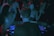 A DJ is performing at a crowded party, standing behind a set of digital turntables and sound equipment. The focus is on the DJ from behind, with a crowd of people visible, immersed in the music and dim lighting, enhanced by colored lights creating an atmospheric ambiance.