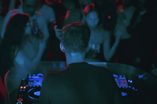 A DJ is performing at a crowded party, standing behind a set of digital turntables and sound equipment. The focus is on the DJ from behind, with a crowd of people visible, immersed in the music and dim lighting, enhanced by colored lights creating an atmospheric ambiance.