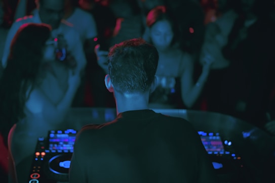 A DJ is performing at a crowded party, standing behind a set of digital turntables and sound equipment. The focus is on the DJ from behind, with a crowd of people visible, immersed in the music and dim lighting, enhanced by colored lights creating an atmospheric ambiance.