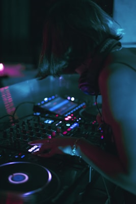 A person is engaged in DJing with a professional setup that includes turntables and a mixer. The dim lighting casts a neon glow, creating an atmospheric scene likely set in a club or music venue. The person's head is slightly bowed, focusing intently on the equipment.