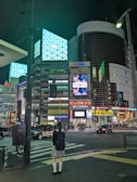 A city street scene at night featuring tall buildings illuminated with bright advertisements and digital screens. There is a girl in a uniform standing at a crosswalk, waiting to cross the street.