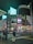 A city street scene at night featuring tall buildings illuminated with bright advertisements and digital screens. There is a girl in a uniform standing at a crosswalk, waiting to cross the street.