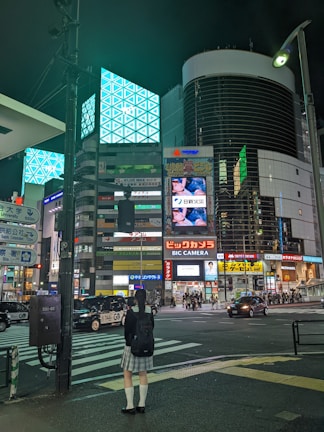 A city street scene at night featuring tall buildings illuminated with bright advertisements and digital screens. There is a girl in a uniform standing at a crosswalk, waiting to cross the street.