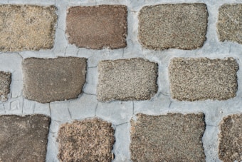 A close-up view of a cobblestone pavement with rectangular stones arranged in a regular pattern. The stones vary in shades of brown and gray, and the gaps between the stones are filled with cement.