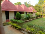 A row of cottages with distinct, reddish-pink, triangular roofs is seen. The cottages have beige walls and green-painted door frames. In front of the cottages, there is a well-maintained garden with various shrubs, bushes, and small trees. A concrete pathway runs alongside the garden.