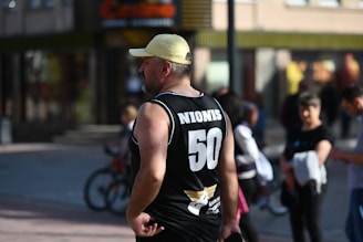 A man wearing a black sports jersey with the number 50 and the name Nionis on the back stands with his hands on his hips. He is wearing a yellow cap and is watching something out of view. In the background, people are walking, and a bicycle is partially visible.