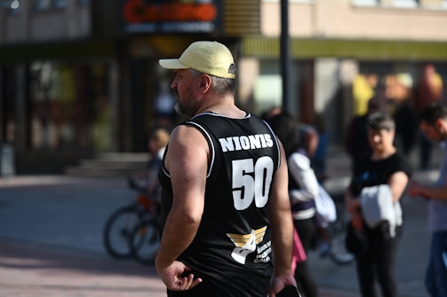 A man wearing a black sports jersey with the number 50 and the name Nionis on the back stands with his hands on his hips. He is wearing a yellow cap and is watching something out of view. In the background, people are walking, and a bicycle is partially visible.