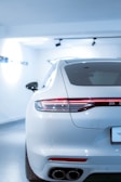 A sleek, white luxury car is parked in a brightly lit showroom. The focus is on the rear of the car, showcasing its modern design, including the streamlined taillights and dual exhaust pipes. The environment is pristine and well-lit, emphasizing the car's polished exterior.