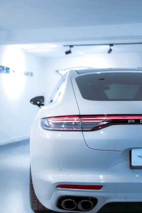 A sleek, white luxury car is parked in a brightly lit showroom. The focus is on the rear of the car, showcasing its modern design, including the streamlined taillights and dual exhaust pipes. The environment is pristine and well-lit, emphasizing the car's polished exterior.
