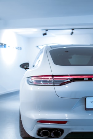 A sleek, white luxury car is parked in a brightly lit showroom. The focus is on the rear of the car, showcasing its modern design, including the streamlined taillights and dual exhaust pipes. The environment is pristine and well-lit, emphasizing the car's polished exterior.