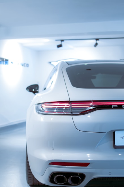 A sleek, white luxury car is parked in a brightly lit showroom. The focus is on the rear of the car, showcasing its modern design, including the streamlined taillights and dual exhaust pipes. The environment is pristine and well-lit, emphasizing the car's polished exterior.
