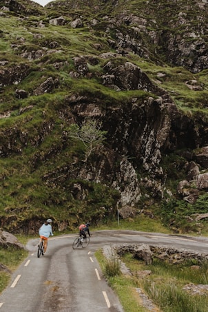 Two cyclists are riding along a narrow, winding road surrounded by lush green hills and rocky terrain. The road curves sharply, adding a sense of adventure and challenge to their journey. The natural landscape is rugged and picturesque, with grass and rocks covering the hillside.