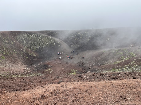 A group of people are scattered across a large volcanic crater landscape. The ground consists of reddish-brown soil mixed with patches of green vegetation. Mist or smoke partially obscures the area, creating a cloudy, mysterious atmosphere.
