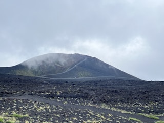 A volcanic landscape with a barren, rocky ground covered in dark ash. A conical volcano rises in the background, partially shrouded in mist or fog. Sparse patches of grass or vegetation are visible, adding small touches of green to the scene. The sky is overcast with a mix of grey clouds.