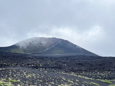 A volcanic landscape with a barren, rocky ground covered in dark ash. A conical volcano rises in the background, partially shrouded in mist or fog. Sparse patches of grass or vegetation are visible, adding small touches of green to the scene. The sky is overcast with a mix of grey clouds.