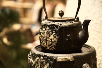 An ornate cast iron teapot with intricate engravings of figures dressed in traditional clothing, highlighting detailed craftsmanship. It is placed on a similarly styled base with a blurred background that suggests an indoor setting.