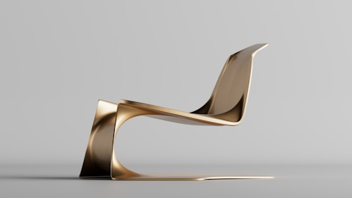 A modern, sculptural chair with a sleek, continuous design crafted from a metallic material, likely gold or brass in color. It has a futuristic aesthetic and sits on a smooth surface with a seamless, flowing structure that extends from the base to the backrest.