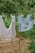 Two light-colored sheets are hanging on a clothesline in a lush, green garden. An old, rusty bed frame is positioned nearby, set among grass and straw-covered ground. Trees with dense foliage surround the area, creating a serene and rustic outdoor setting.