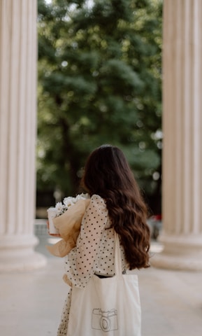 A woman with long, wavy brown hair stands between two columns, holding a bouquet of white flowers wrapped in brown paper and a coffee cup. She wears a white polka-dotted dress and carries a white tote bag with a camera illustration. The background features blurry green foliage.