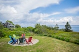 A tranquil coastal scene with a charming wooden house nestled among green trees and shrubbery. There is a grassy area with a circle of colorful Adirondack chairs overlooking the ocean. The sky is partly cloudy, adding to the serene atmosphere of the landscape.