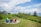 A tranquil coastal scene with a charming wooden house nestled among green trees and shrubbery. There is a grassy area with a circle of colorful Adirondack chairs overlooking the ocean. The sky is partly cloudy, adding to the serene atmosphere of the landscape.