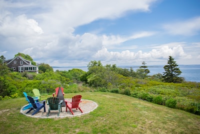 A tranquil coastal scene with a charming wooden house nestled among green trees and shrubbery. There is a grassy area with a circle of colorful Adirondack chairs overlooking the ocean. The sky is partly cloudy, adding to the serene atmosphere of the landscape.