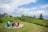 A tranquil coastal scene with a charming wooden house nestled among green trees and shrubbery. There is a grassy area with a circle of colorful Adirondack chairs overlooking the ocean. The sky is partly cloudy, adding to the serene atmosphere of the landscape.