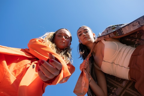 Two individuals are looking down at the camera, wearing fashion-forward outfits. The person on the left has long, wavy hair and is dressed in a vibrant orange suit. The person on the right is wearing a patterned coat over a white top. Both have serious expressions and the bright blue sky serves as a backdrop, enhancing the bold colors of their clothing.