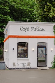 A quaint café with a white exterior and an orange trim, featuring a single window and a glass door. In front of the café, there are two metal chairs and a small round table on a paved surface. The background consists of lush green foliage from nearby trees.