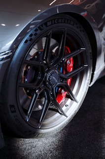 A close-up view of a high-performance car wheel featuring a sleek black alloy rim with a prominent Michelin tire. The wheel reveals a detailed red brake caliper inside, suggesting a sporty and powerful automotive design.