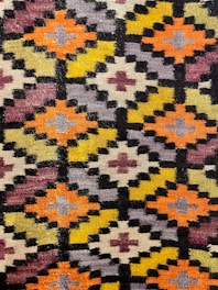 A detailed view of a geometric patterned textile with repetitive diamond shapes arranged in a grid. The pattern includes colors like orange, yellow, purple, and black with smaller crosses inside each diamond. The textile has a slightly fuzzy texture, indicating a woven or knitted fabric.