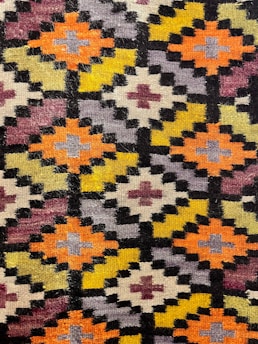 A detailed view of a geometric patterned textile with repetitive diamond shapes arranged in a grid. The pattern includes colors like orange, yellow, purple, and black with smaller crosses inside each diamond. The textile has a slightly fuzzy texture, indicating a woven or knitted fabric.