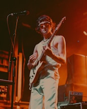 A musician wearing sunglasses and a sleeveless top plays an electric guitar on stage. The setting is dimly lit with warm orange and yellow stage lights creating an atmospheric effect. A microphone stands nearby, and there appear to be speakers and other equipment in the background.
