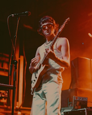 A musician wearing sunglasses and a sleeveless top plays an electric guitar on stage. The setting is dimly lit with warm orange and yellow stage lights creating an atmospheric effect. A microphone stands nearby, and there appear to be speakers and other equipment in the background.