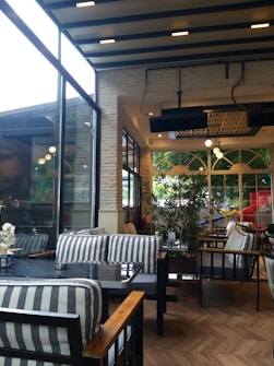 An interior view of a cozy caf&eacute; with a modern design. There are striped cushioned chairs around glass-top tables. The space is well-lit with large windows letting in natural light. Some potted plants are placed strategically to enhance the ambiance. The ceiling has exposed beams and contemporary lighting fixtures.
