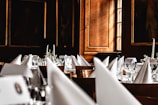 A luxurious dining setup featuring elegantly folded white napkins, sparkling glassware, and floral centerpieces arranged on round tables. The room is adorned with wooden paneling and framed portraits, creating an opulent and classic atmosphere.