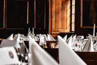 A luxurious dining setup featuring elegantly folded white napkins, sparkling glassware, and floral centerpieces arranged on round tables. The room is adorned with wooden paneling and framed portraits, creating an opulent and classic atmosphere.