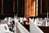 A luxurious dining setup featuring elegantly folded white napkins, sparkling glassware, and floral centerpieces arranged on round tables. The room is adorned with wooden paneling and framed portraits, creating an opulent and classic atmosphere.