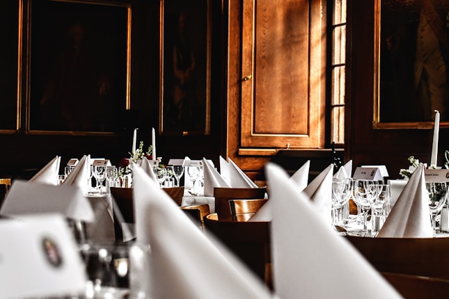 A luxurious dining setup featuring elegantly folded white napkins, sparkling glassware, and floral centerpieces arranged on round tables. The room is adorned with wooden paneling and framed portraits, creating an opulent and classic atmosphere.