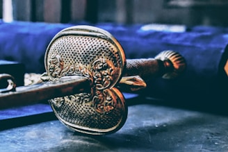 An ornate and intricately designed metal object, possibly a sword hilt or a key, lies in focus against a blurred background. The object features detailed engravings and a textured surface, suggesting craftsmanship and historic significance. The colors are rich and give it a sense of antiquity.