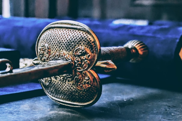 An ornate and intricately designed metal object, possibly a sword hilt or a key, lies in focus against a blurred background. The object features detailed engravings and a textured surface, suggesting craftsmanship and historic significance. The colors are rich and give it a sense of antiquity.
