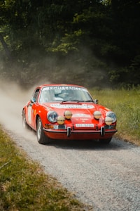A vintage red rally car is driving along a dirt road surrounded by greenery. The car is decorated with various decals and has several additional front lights. A banner with 'Fight Cancer' is displayed across the windshield. Dust is being kicked up behind the car as it races forward.
