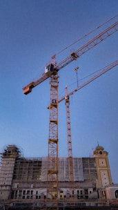 Several construction cranes stand amidst a partially constructed building. The building has scaffolding and netting covering sections of its facade. The sky in the background is clear and blue, indicating daytime.
