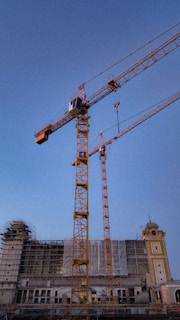 Several construction cranes stand amidst a partially constructed building. The building has scaffolding and netting covering sections of its facade. The sky in the background is clear and blue, indicating daytime.
