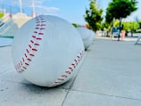 Large sculptural baseballs with red stitching are placed along a sidewalk, with a park-like setting in the background featuring trees and a clear sky.