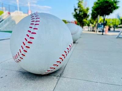 Large sculptural baseballs with red stitching are placed along a sidewalk, with a park-like setting in the background featuring trees and a clear sky.