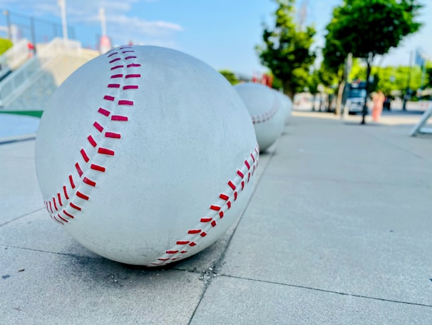 Large sculptural baseballs with red stitching are placed along a sidewalk, with a park-like setting in the background featuring trees and a clear sky.