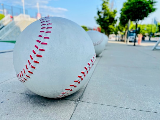 Large sculptural baseballs with red stitching are placed along a sidewalk, with a park-like setting in the background featuring trees and a clear sky.