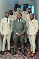 A group of eleven men stand together in a hallway, all dressed in formal attire. The central figure wears a green suit, while the others wear cream-colored suits. They appear confident, with some wearing glasses, and many have boutonnières pinned to their lapels. The hallway has patterned carpeting and light walls.