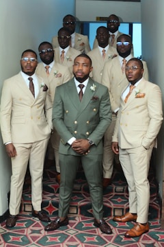 A group of eleven men stand together in a hallway, all dressed in formal attire. The central figure wears a green suit, while the others wear cream-colored suits. They appear confident, with some wearing glasses, and many have boutonnières pinned to their lapels. The hallway has patterned carpeting and light walls.